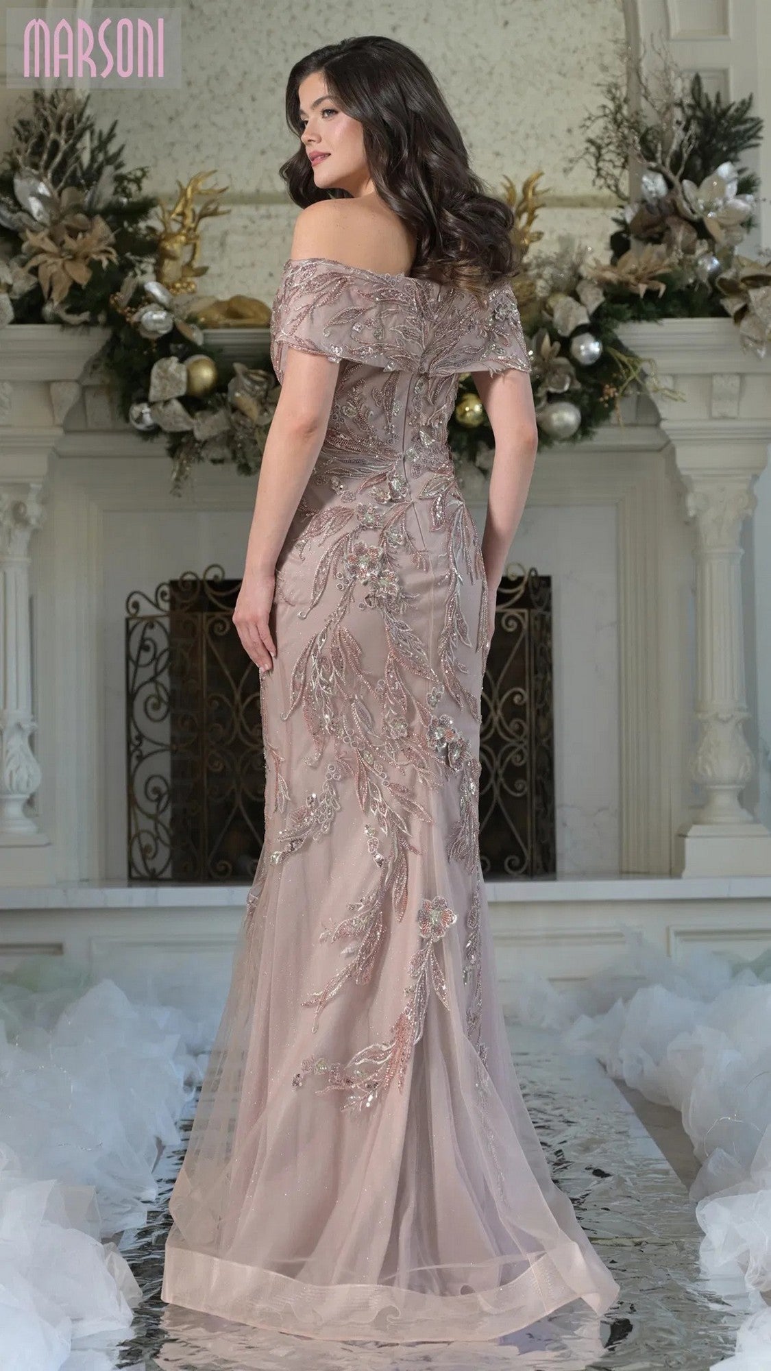 A woman wearing the Marsoni MV1372 long formal dress with a mermaid silhouette and delicate beaded embroidery stands before a decorated fireplace, elegantly glancing over her shoulder.